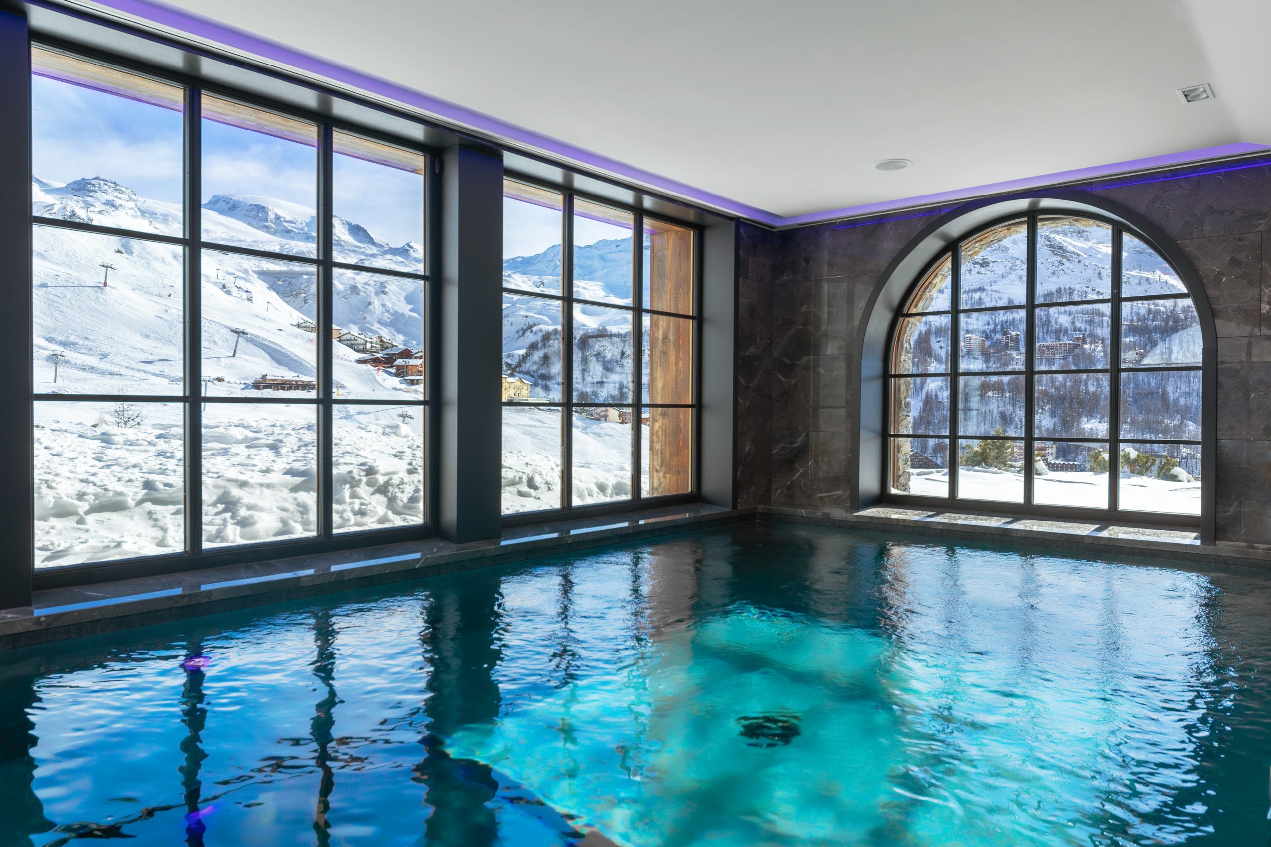 The luxury swimming pool of Chalet La Fenice, a lxuury ski chalet in Cervinia. Outside the tall windows are views of the mountains and the slopes, and the water reflects stylish purple llights.