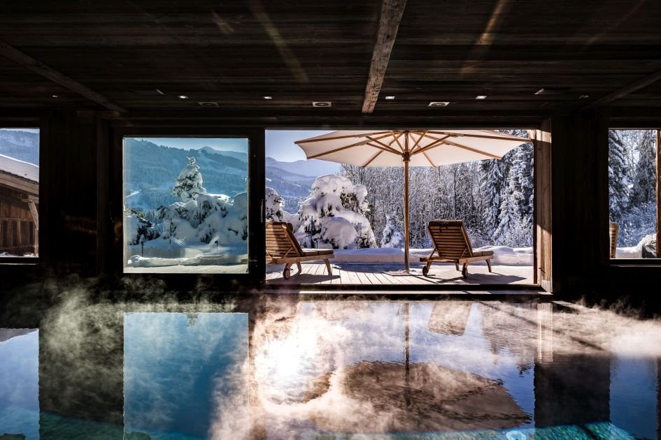 A view of Ultima Megeve's steaming swimming pool with an outdoor terrace and snowy mountain tops. 