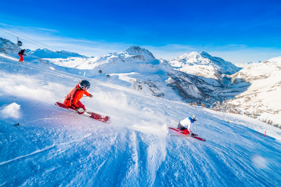 Two skiers on the Piste M slope on the Solaise mountain in Val d'Isère with blue skies and the village in the valley below. 