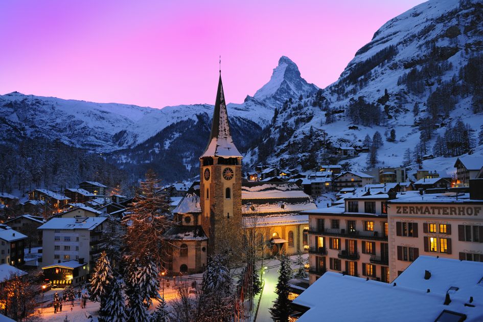The church and surrounding village of Zermatt at sunset, with the Matterhorn in the background against a pink sky. 